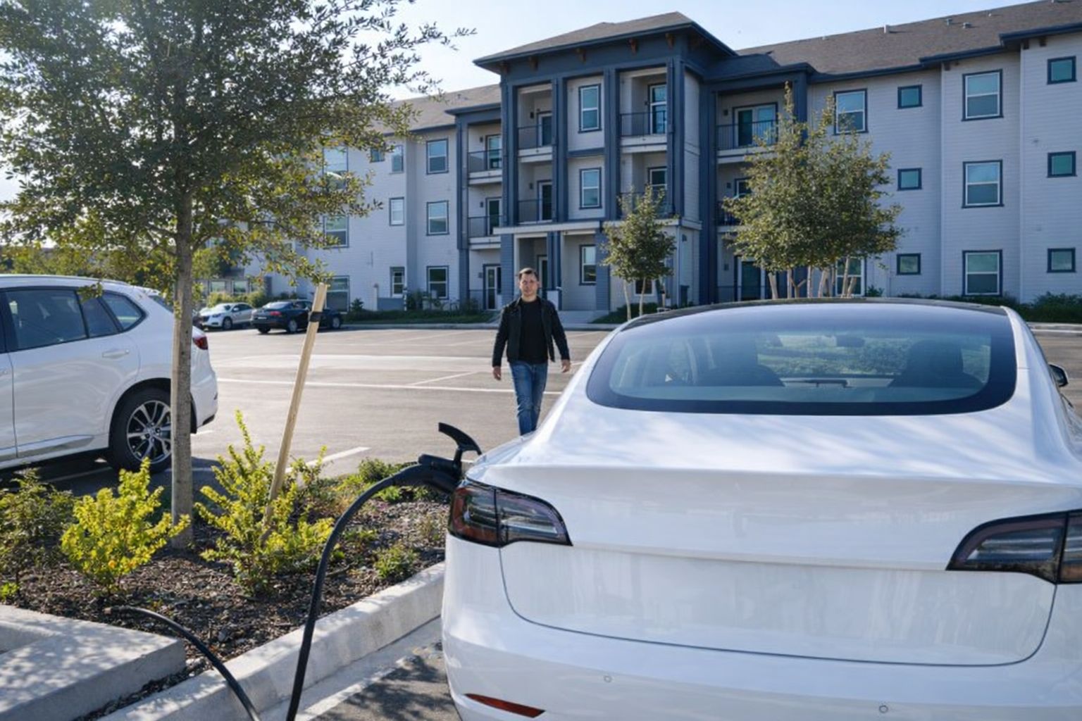 En vit elbil laddas på en parkeringsplats framför ett lägenhetshus.